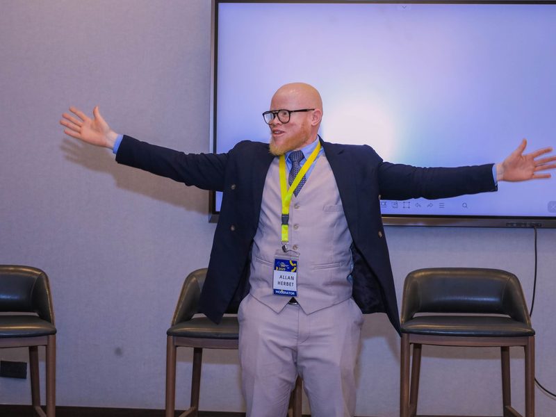 Hosting on stage, arms open, addressing a conference room